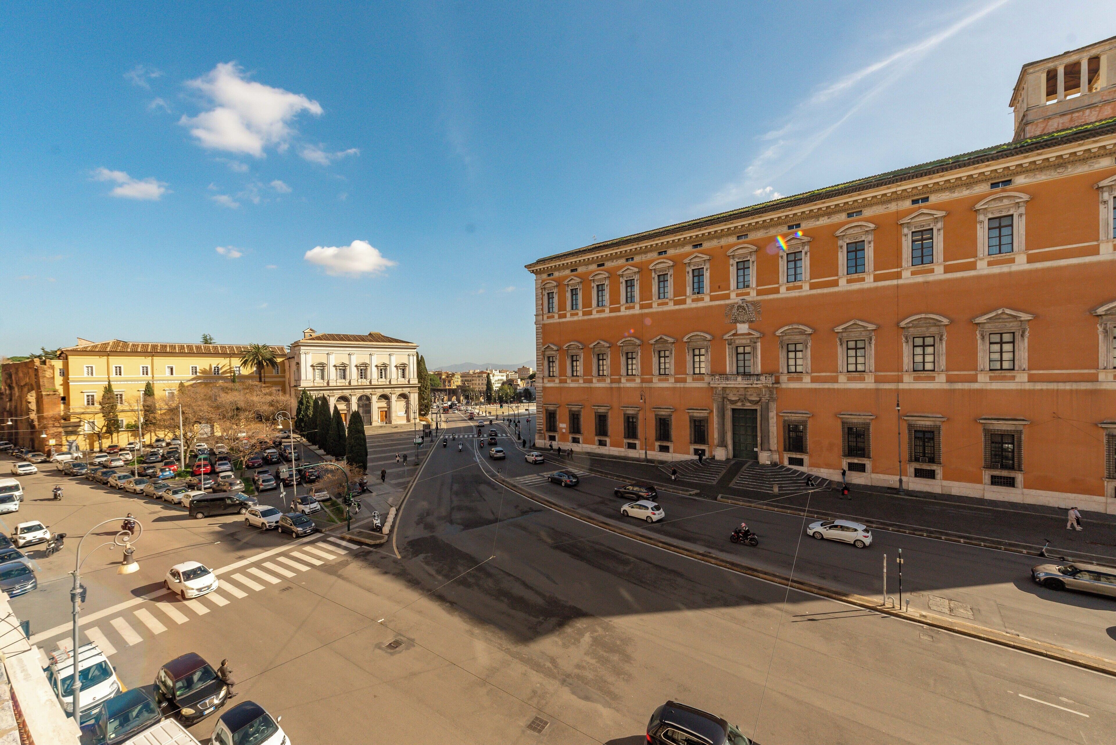 A wide street with buildings on either side, including a prominent red building with classical architecture.