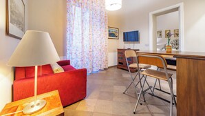Ein Wohnzimmer mit einem roten Sofa, einem Holztisch mit einer Lampe, einem Schreibtisch mit Stuhl, einem Fernseher und einem Fenster mit floralen Vorhängen.