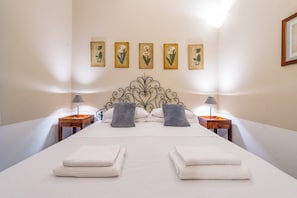 Un lit bien fait, avec du linge de lit blanc et deux oreillers, une tête de lit en bois et une série d’œuvres d’art florales encadrées accrochées au mur.