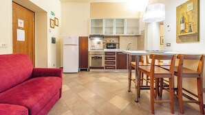 Eine kompakte Küche mit Kühlschrank, Backofen, Mikrowelle und Holzschränken. Im Wohnbereich befindet sich ein Esstisch mit Stühlen sowie eine rote Couch.