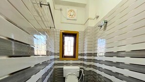 Un baño estrecho, con baldosas, que incluye un inodoro, un ventanal con marco de madera y un ventilador fijado a la pared.