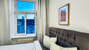 Un dormitorio con cama, ventana con cortinas, radiador, cabecera tapizada en tono café y un cuadro en la pared.