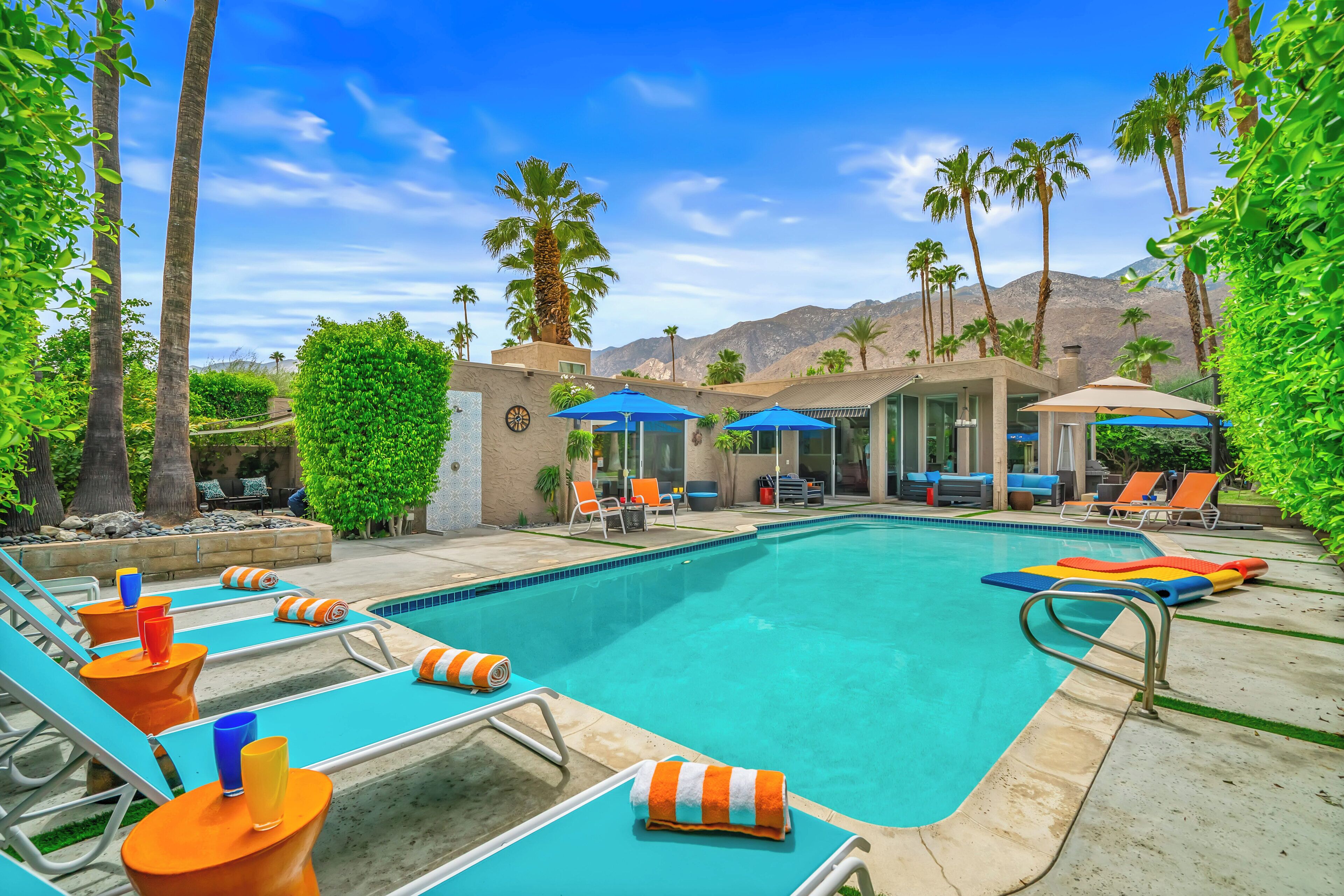 A swimming pool with lounge chairs and umbrellas, surrounded by palm trees and mountains in the background.