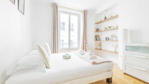 Une chambre à coucher avec un lit, une fenêtre avec des rideaux, une table de chevet avec une lampe et une étagère avec des objets décoratifs.