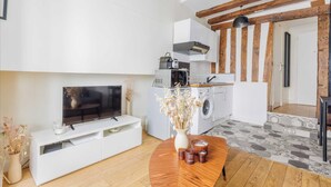 Un salon moderne doté d’un téléviseur à écran plat, d’un meuble de rangement blanc, d’une table basse en bois agrémentée d’objets décoratifs, et d’un coin cuisine comprenant un lave-linge et un four à micro-ondes.