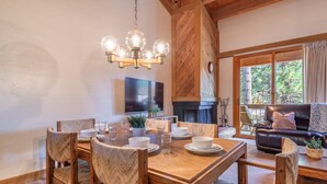 Un comedor con una mesa de madera, sillas y una araña. Se ve una chimenea, un sofá y un televisor al fondo.