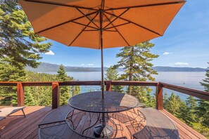 Una terraza de madera con una mesa redonda y una sombrilla, con vistas a un lago y montañas.