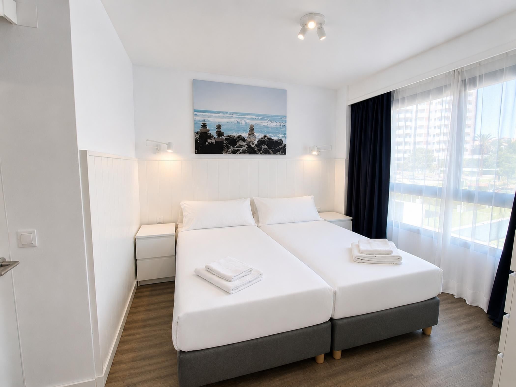 Un lit double avec deux oreillers et une serviette pliée, une table de chevet, une lampe fixée au mur, un tableau représentant une côte rocheuse et une fenêtre avec des rideaux légers.