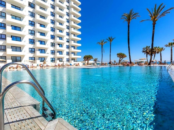 A swimming pool with clear blue water, surrounded by palm trees and a white building with balconies.