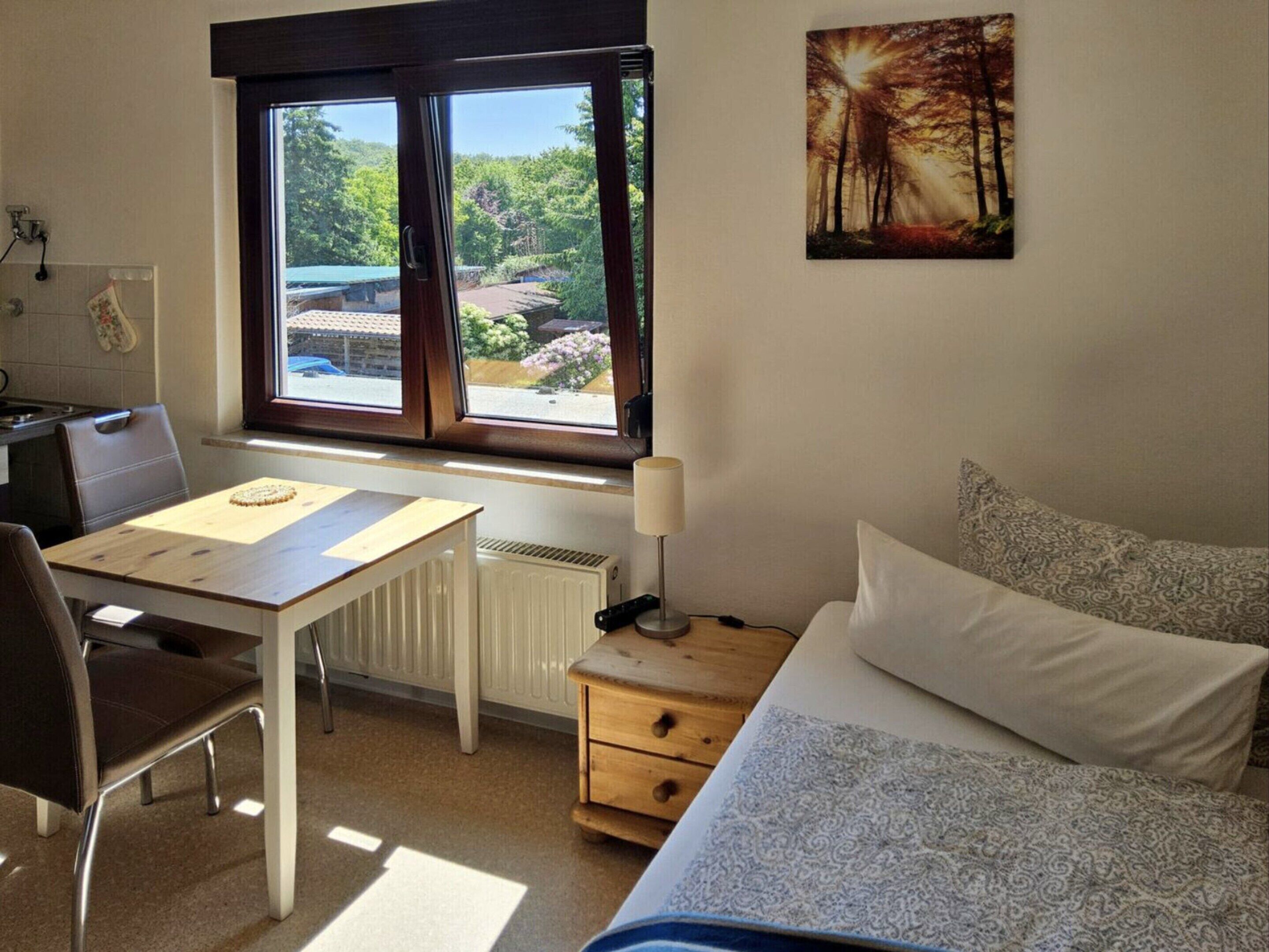 A bedroom with a bed, a desk, a chair, a lamp, and a window with a view of greenery.