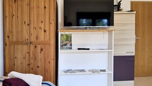 Un dormitorio con un armario de madera, un televisor sobre una estantería blanca y una cama con una manta a rayas.