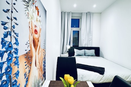A bedroom with a bed, a nightstand, a lamp, a window with curtains, and a wall with a floral mural.