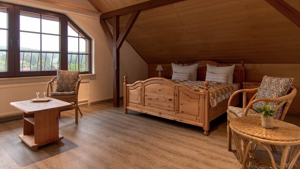 Une chambre mansardée chaleureuse, avec un lit en bois, deux fauteuils en osier, une petite table et de grandes fenêtres donnant sur un paysage verdoyant.