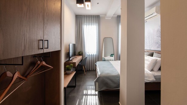 Ein modernes Hotelzimmer mit einem Holzschrank, einem Schreibtisch mit Stuhl, einem Bett mit weißer und grauer Bettwäsche und einem großen Spiegel.