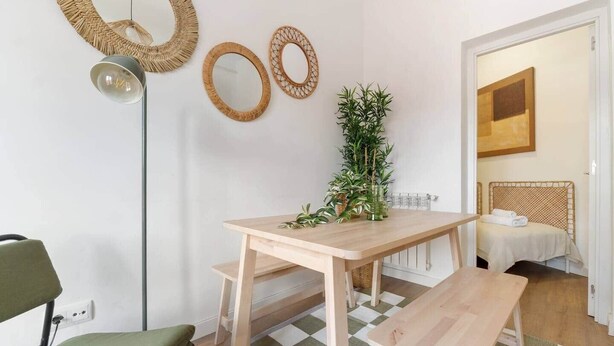 A room with a wooden table, a green chair, a floor lamp, and a bed with a woven headboard.