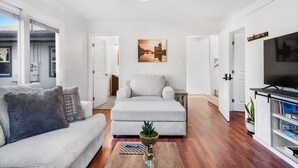 A modern living room with a grey sofa, a matching ottoman, a coffee table with a plant, a flat-screen TV, and a painting on the wall.