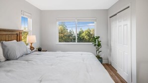 A bedroom with a large bed, a nightstand with a lamp, a window with a view of trees, and a door. - Pet-Friendly, Stylish Home w/ Backyard Oasis (Santa Maria)