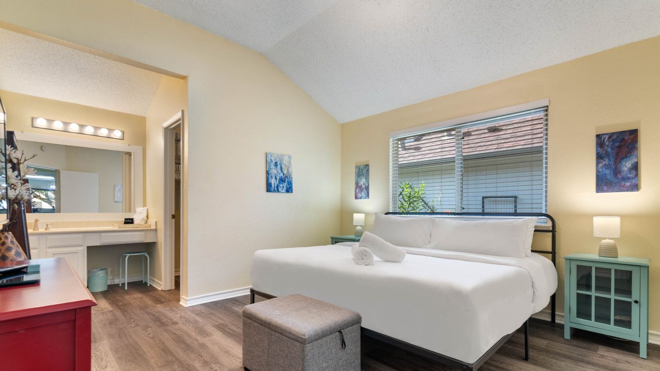 A bedroom with a bed, a vanity with a mirror, and a window with blinds.