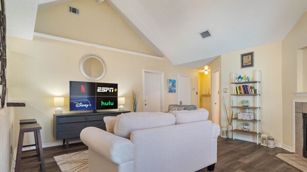 A living room with a flat-screen TV displaying streaming services, a sofa, a coffee table, a bookshelf, and a fireplace.