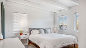 A bedroom with a large bed, a bedside table, a nightstand, a potted plant, and a window with blinds.
