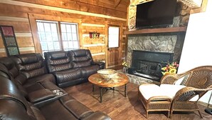 Ein rustikaler Innenraum einer Blockhütte mit Ledersofa, Korbstühlen, Kamin und Holzwänden.