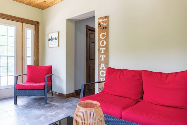 Une pièce avec un canapé rouge, un fauteuil en osier avec des coussins rouges, une lampe en osier et une pancarte indiquant : « Bienvenue au COTTAGE ».