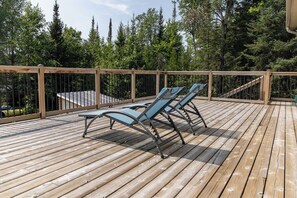 Um deck de madeira com espreguiçadeiras e guarda-corpo com vista para uma área arborizada.