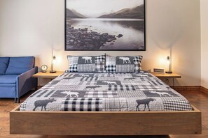 Ein Schlafzimmer mit einem Bett, einem blauen Sessel, einem Nachttisch mit Lampe und einem gerahmten Bild an der Wand.