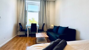 A compact hotel room with a bed, a small table, a sofa, a chair, and a window with curtains. - Convenient Room in Shared Apt | Walk to Kaufpark (Vienna)