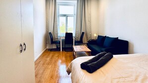 A hotel room with a bed, a desk, two chairs, a sofa, and a window with curtains. - Convenient Room in Shared Apt | Walk to Kaufpark (Vienna)