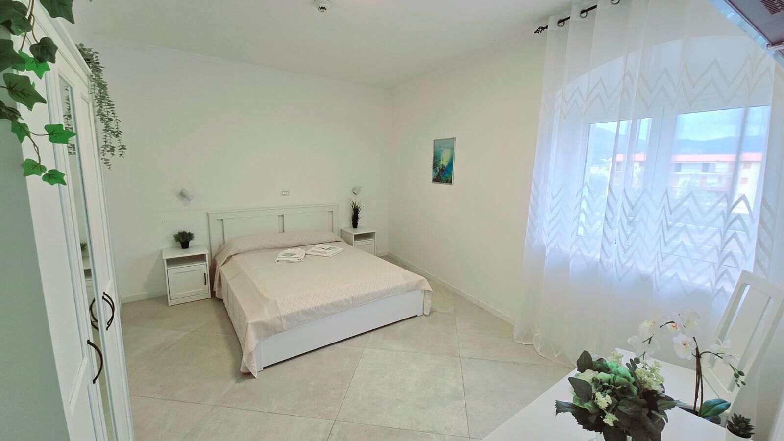 A bedroom with a bed, bedside tables, a nightstand, a window with sheer curtains, and a view of buildings outside.