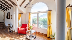 A spacious room with a red chair, a small table with a plant, a large window with yellow curtains, and a view of greenery outside.