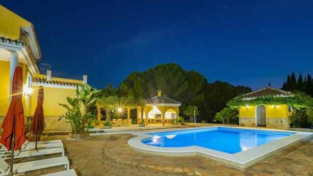 A swimming pool with a tiled deck, a covered seating area, and a building with yellow walls and a tiled roof.