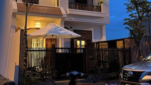 A two-story house with a balcony, a scooter parked in front, and a car parked on the street.