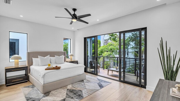Un dormitorio moderno con una cama grande, un ventilador de techo, una mesita de noche con lámpara y vistas a un balcón con mobiliario exterior.