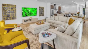 Una sala moderna con un sofá, sillones y una mesa de centro. Un televisor está montado en la pared, y se ve una cocina al fondo.