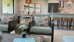 A modern living room with a sofa set, a coffee table with a book, and a kitchen area with a refrigerator and bar stools. - Ma'alaea Surf, Unit A4-2 BR Ground Floor Beachfront Condo, AC, Wi-Fi, Pool (Kihei)