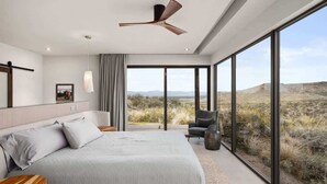 A modern bedroom with a large bed, a ceiling fan, a view of the desert through large windows, and a small seating area. - Paradise Canyon-Infinity Pool-Mnt Views-Estate 3 (Fort Mcdowell)