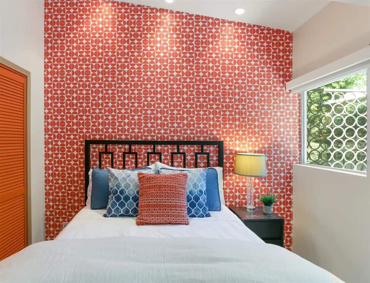 A bedroom with a patterned wallpaper, a bed with multiple pillows, a bedside table with a lamp, and a window with a view.