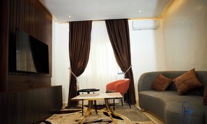 A modern hotel room with a flat-screen TV mounted on the wall, a grey sofa with brown cushions, a small round table, and a patterned rug. - Harzock hotel (Abuja)