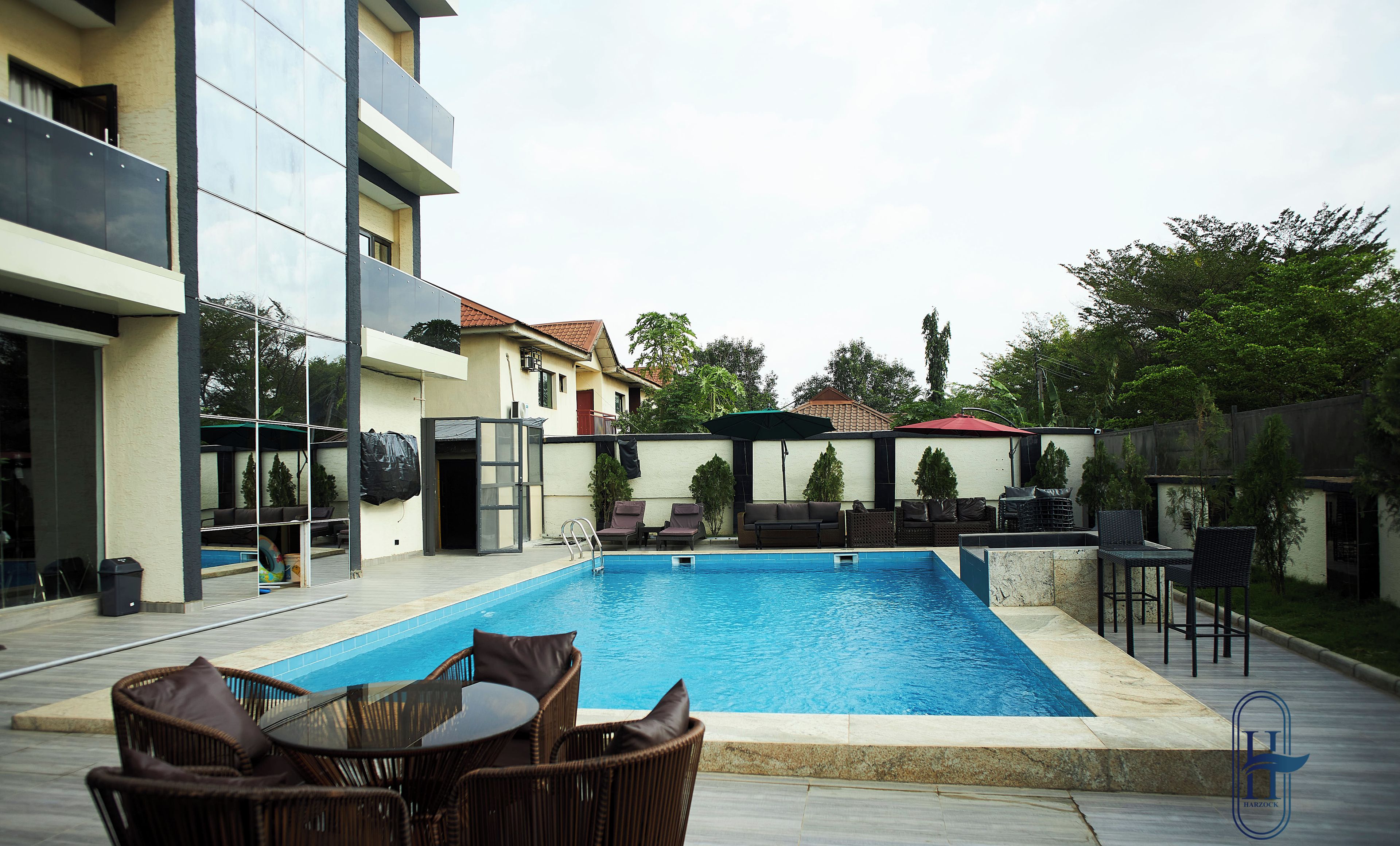 A swimming pool with lounge chairs and a table, surrounded by a modern building with balconies and a fenced area with outdoor furniture.