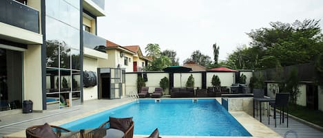 A swimming pool with lounge chairs and a table, surrounded by a modern building with balconies and a fenced area with outdoor furniture.