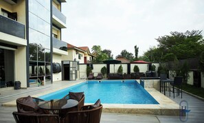A swimming pool with lounge chairs and a table, surrounded by a modern building with balconies and a fenced area with outdoor furniture. - Harzock hotel (Abuja)