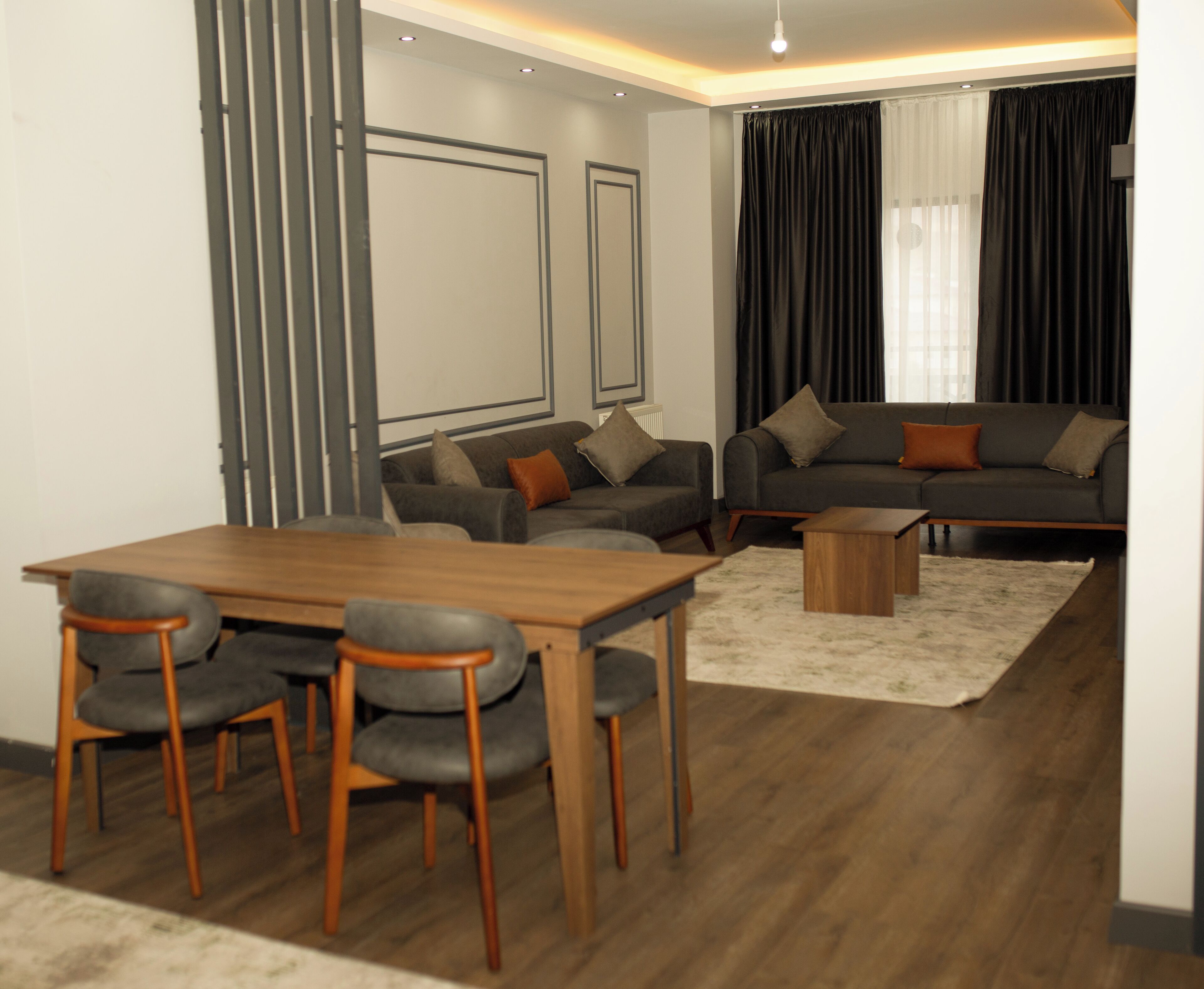 Un salon moderne avec une table et des chaises de salle à manger en bois, un ensemble de canapés gris et une grande fenêtre avec des rideaux foncés.