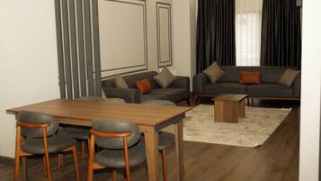 Un salon moderne avec une table et des chaises de salle à manger en bois, un ensemble de canapés gris et une grande fenêtre avec des rideaux foncés.