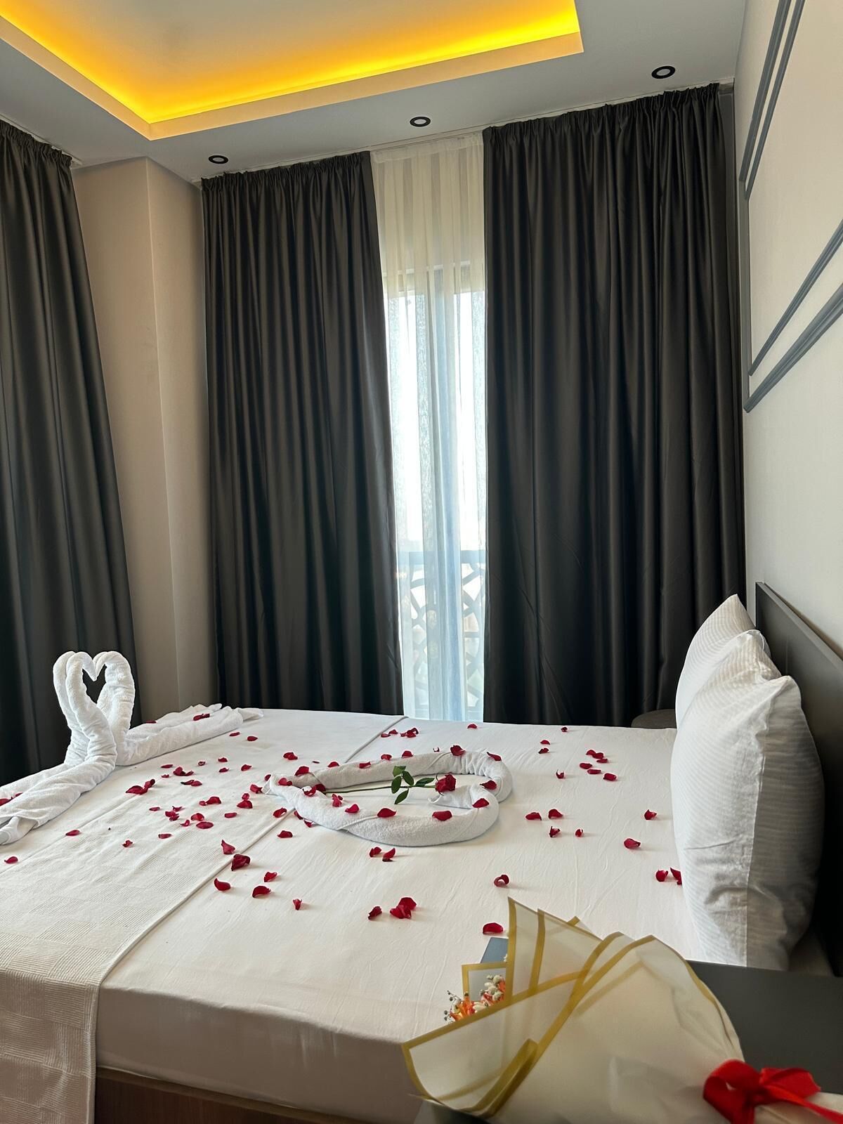 A hotel room with a neatly made bed, a heart-shaped towel arrangement, and rose petals scattered on the bed.