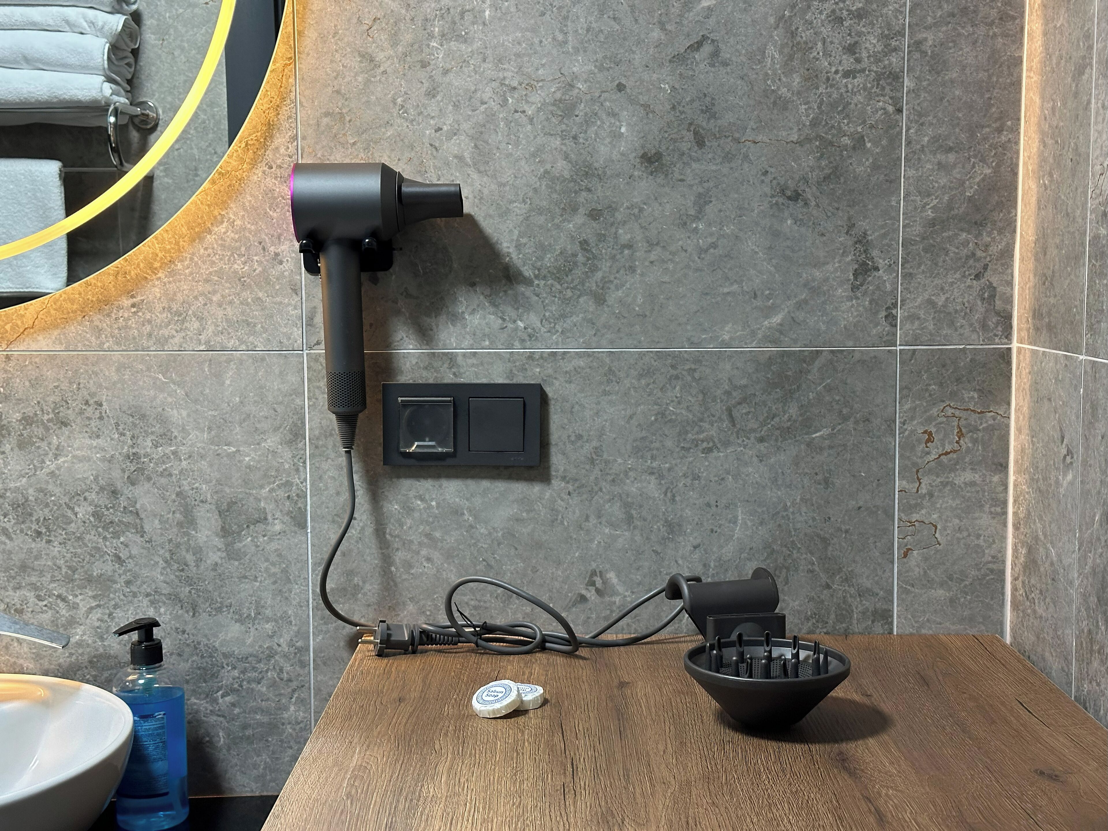 Un baño con un secador de pelo montado en la pared, dispensador de jabón y un recipiente con artículos de aseo sobre una superficie de madera.