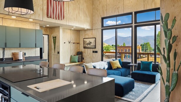 A modern kitchen with a central island, dining area with blue sofas, and large windows offering a mountain view.