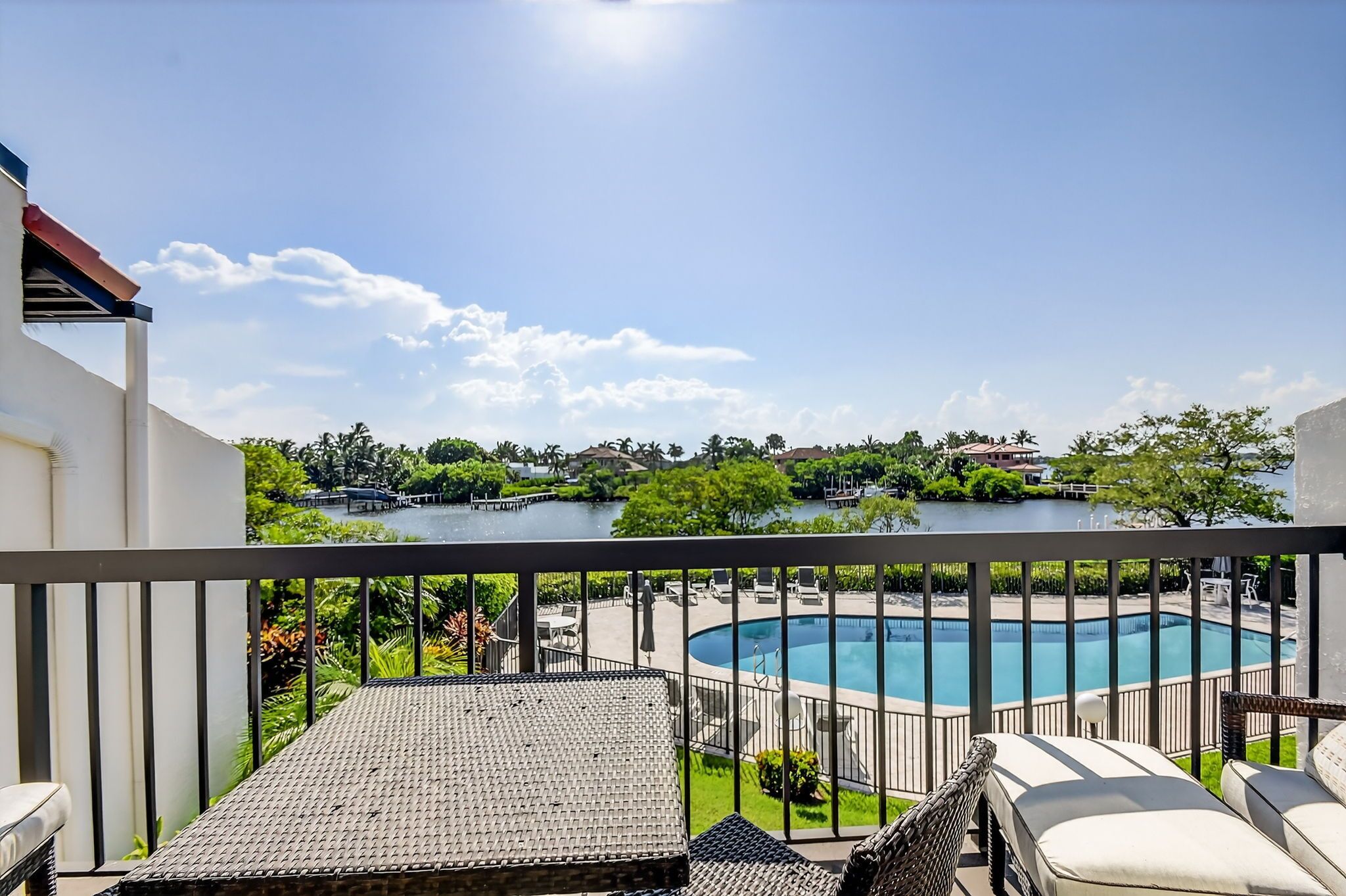 A balcony with a view of a swimming pool, lush greenery, and a body of water.
