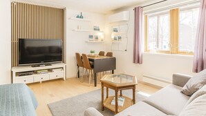 Een moderne woonkamer met een flatscreen-tv, een eethoek met een tafel en stoelen, en een groot raam met gordijnen.